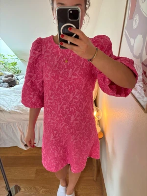 Rosa kort klänning med puffärmar - Superfin rosa kort klänning med strukturmönster och stora puffärmar. Klänningen har rund halsringning och en rak passform som ger en trendig look. Perfekt för dig som vill sticka ut med färg och volym. Materialet känns lite tjockare och har en snygg textur.