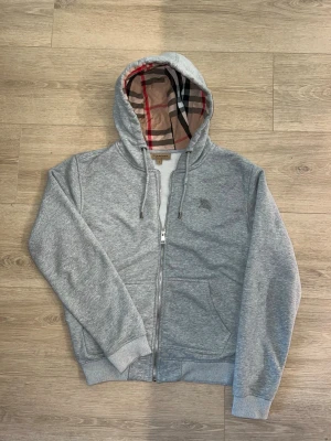 Burberry Hoodie - Snygg grå hoodie med dragkedja från Burberry. Klassisk huva med det ikoniska rutmönstret på insidan, ribbade muddar och fickor framtill. Perfekt för en chill och stilren look. Materialet är mjuk bomullsblandning.