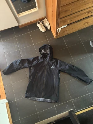 Svart regnjacka från Arcteryx - Snygg svart regnjacka från Arc'teryx med huva och hel dragkedja framtill. Jackan har två snedställda fickor med dragkedja och är tillverkad i ett vattentätt material. Insidan är ljusgrå och har en innerficka med Arc'teryx-logga. Perfekt för regniga dagar.                   Viktigt: Står xxl men är för liten för mig så skulle säga den passar xl. 