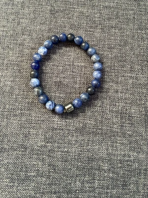 Blått pärlarmband med stendetaljer - Snyggt armband med runda blå och grå pärlor i olika nyanser, trädda på elastisk tråd. Armbandet har en metallisk kubformad detalj som accent. Perfekt accessoar för att lyfta din stil.