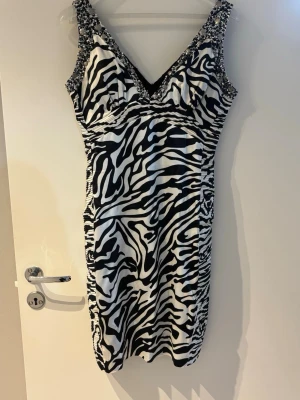 Zebramönstrad kort klänning med pärlor - Snygg kort klänning med zebramönster i svart och vitt. Klänningen har djup v-ringning och breda axelband dekorerade med pärlor. Figurnära passform som ger en cool och trendig look.