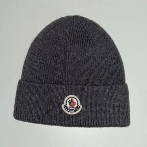 Grå Moncler mössa i ull - Säljer en mörkgrå ribbstickad mössa från Moncler med uppvikt kant och klassisk Moncler-logga framtill. Mössan är tillverkad i mjuk ull och har en enkel, stilren design som passar perfekt till kalla dagar.