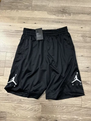 Svarta Jordan Air shorts - Svarta sportiga shorts från Jordan med vit Air-logga på båda benen. Tillverkade i ett lätt och ventilerande polyestermaterial med elastisk midja för skön passform. Perfekta för träning eller en avslappnad stil.