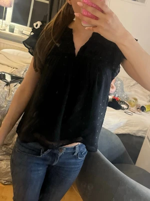 Svart blus med volangärmar - Snygg svart blus med volangärmar och lätt v-ringning. Blusen har ett diskret prickigt mönster i tyget och är tillverkad i ett tunt, luftigt material som ger en avslappnad look. Perfekt att matcha med jeans eller kjol för en trendig outfit. Storlek 158-164