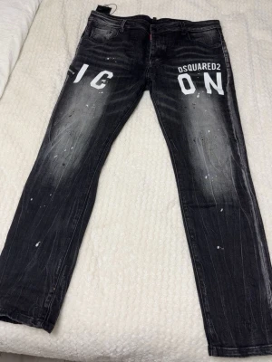 Svarta Dsquared2 ICON jeans - Snygga svarta jeans från Dsquared2 med vit 'ICON'-text framtill. Jeansen har en slim fit-modell, slitna detaljer och vita färgstänk för en trendig look. Klassisk femficksdesign och tydliga tvättade partier ger extra edge. OBS: storlek 54/32