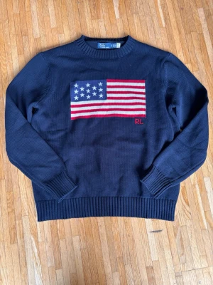  Polo Ralph Lauren flag knitt - Mörkblå stickad tröja från Polo Ralph Lauren med amerikansk flagga på bröstet och RL-broderi. Tröjan har rund hals, ribbstickade muddar och är gjord i bomull. Perfekt för dig som gillar klassisk och stilren design. OBS storlek xl men sitter som l och är slimfit. Skriv privat innan ni köper 