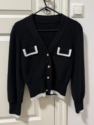 Svart kofta med vita detaljer - Stilren svart kofta med v-ringning, dekorativa fickor med vita kanter och pärlknappar framtill. Ribbstickade muddar och nederkant med vit kontrast. 