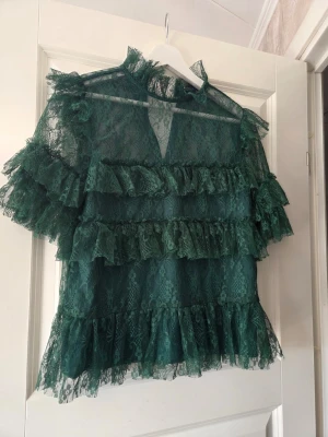 Grön spetsblus med volanger - Snygg grön blus i transparent spets med flera volangdetaljer och hög krage. Blusen har korta ärmar och knäppning i nacken. Perfekt för dig som vill sticka ut med en trendig och romantisk look.