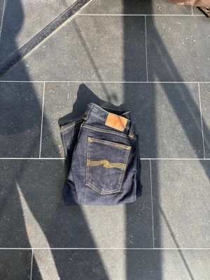 Mörkblå jeans från Nudie Jeans - Säljer ett par mörkblå jeans från Nudie Jeans med klassisk femficksmodell och raka ben. Jeansen har kontrastsömmar i gult och en läderpatch bak i midjan. Tillverkade i kraftig denim som ger en snygg och tidlös look. W30 L34