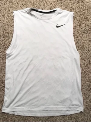 Vit ärmlös linnetopp från Nike - Snygg vit ärmlös linnetopp från Nike med svart logga på bröstet. Tillverkad i ett lätt och svalt material som passar perfekt för träning. Klassisk och sportig design med rund halsringning.