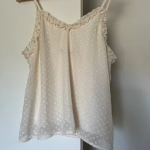 Prickig topp med volang - Supersöt ljusbeige linnetopp med små prickar och volangdetaljer längs axelband och urringning. Tunt, luftigt material och smala axelband gör den perfekt för varma dagar. Enkel att styla med jeans eller kjol.