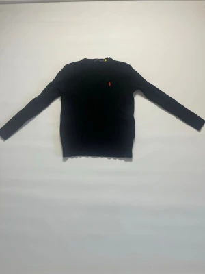 Svart kabel stickad tröja från Ralph Lauren storlek M - Snygg Svart kabelstickad tröja från Ralph Lauren med klassisk röd logga broderad på bröstet. Tröjan har rund halsringning, långa ärmar och ribbade muddar vid ärmslut och nederkant. Perfekt till höst och vinter för en stilren look. Tröjan är i storlek Medium och är aldrig buren