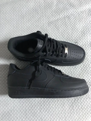 Nike Air Force 1 Low helsvarta sneakers - Säljer ett par klassiska Nike Air Force 1 Low sneakers i helsvart läder. Skorna har perforerad tå, svart sula och matchande svarta skosnören med en metallbricka framtill. Perfekta för en clean och stilren look.