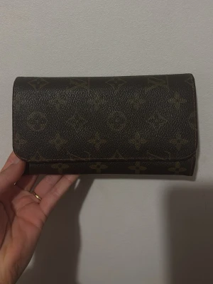 En brun praktisk plånbok - Snygg plånbok i klassiskt brunt monogrammönster. Plånboken är rektangulär och tillverkad i tåligt läder med logga över hela ytan. Perfekt för dig som gillar lyxiga accessoarer och vill ha något stilrent. Den är otroligt praktisk och simpel att bära runt med. #brownwallet#style#vintage