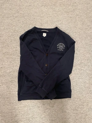 Mörkblå cardigan från Boomerang - Mörkblå cardigan från Boomerang med broderad logotyp och text på bröstet. Tröjan har v-ringning, knappar framtill och två fickor. Materialet är mjukt och bekvämt, perfekt för att slänga på sig när det är lite kyligt.