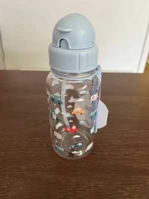 Vattenflaska med bilar och blått lock - Säljer en genomskinlig vattenflaska i plast med blått lock och sugrör. Flaskan är dekorerad med små bilar och moln i olika färger. Perfekt att ha med sig till skolan eller träningen. Praktiskt handtag på sidan för enkel transport.
