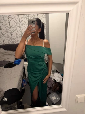 Grön långklänning med slits - Elegant grön långklänning med offshoulder och smala axelband. Klänningen har en snygg slits framtill och en draperad detalj vid midjan. Perfekt för dig som vill sticka ut med en stilren och modern look.