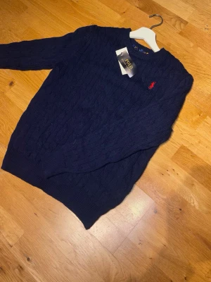 Mörkblå kabelstickad tröja Polo Ralph Lauren - Mörkblå kabelstickad tröja från Polo Ralph Lauren med rund halsringning och den klassiska röda logotypen broderad på bröstet. Tröjan har ribbade muddar vid ärmslut och nederkant. Perfekt för dig som gillar stilren och tidlös design.