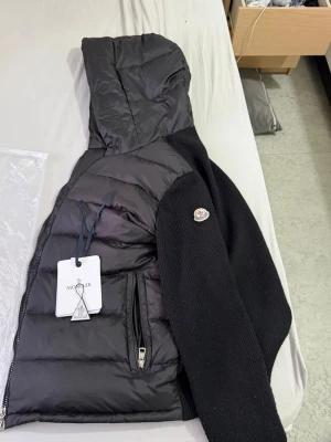 Svart dunjacka med stickade ärmar Moncler - Snygg svart dunjacka från Moncler med stickade ärmar och huva. Jackan har dragkedja framtill, två fickor med dragkedja och Moncler-logga på ärmen. Kroppen är i glansigt dunmaterial medan ärmarna är i mjuk stickad ull.
