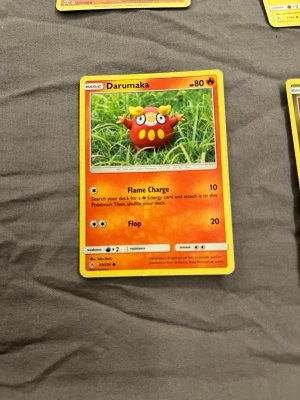 Pokémonkort Darumaka - Säljer ett Pokémonkort med Darumaka-motiv. Kortet har en färgstark design med Darumaka i rött och gult mot en grön bakgrund. Kortet är gjort av laminerad kartong och har attacker som Flame Charge och Flop. Perfekt för samlare eller Pokémonfans.