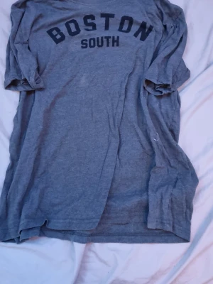 Grå t-shirt med BOSTON-tryck SOC - Grå t-shirt från SOC med svart text 'BOSTON SOUTH' på bröstet. Klassisk rund hals och avslappnad passform. Perfekt för en chill look och enkel att matcha med jeans eller shorts.