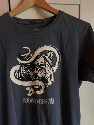 Mörkblå Roberto Cavalli t-shirt - Cool mörkblå t-shirt från Roberto Cavalli med ett stort tryck av en tiger och en orm på bröstet. Rund halsringning och korta ärmar. Materialet är mjukt och bekvämt, perfekt för dig som gillar unika designer.
