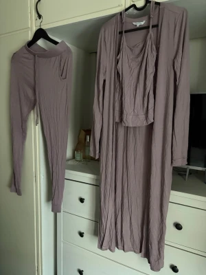 Lila lounge-set  - Säljer ett lila lounge-set med långärmad cardigan med knappar, matchande linne med smala axelband och ett par mjuka byxor med dragsko i midjan. Setet är gjort i ett tunt, stretchigt material som känns skönt mot huden. Perfekt för en avslappnad look.