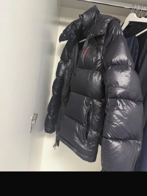 Svart pufferjacka från Ralph Lauren - Snygg svart pufferjacka från Ralph Lauren med glansig finish och röd broderad logga på bröstet. Jackan har hög krage, dragkedjor på fickorna och en klassisk quiltad design. Perfekt för kalla vinterdagar och riktigt varm. JACKAN ÄR ÄKTA 