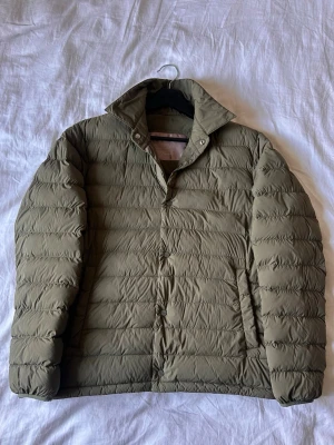 Olivgrön dunjacka från Herno - Snygg olivgrön dunjacka från Herno med klassisk krage och diskreta sidofickor. Jackan är quiltad med horisontella sömmar och har tryckknappar framtill. Perfekt för vårdagar och enkel att matcha med olika outfits. Storlek 48 vilket motsvarar S/M