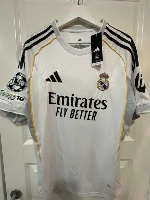 Real Madrid matchtröja Adidas vit - Snygg vit Real Madrid matchtröja från Adidas med svarta och gula detaljer. Tröjan har klubbmärke, Adidas-logga och sponsortryck på bröstet samt patchar på ärmarna. Tillverkad i lätt och ventilerande material, perfekt för fotboll eller träning.