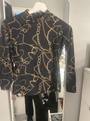 Svart långärmad topp från CRW - Svart långärmad topp från CRW med coolt kedjemönster i guld. Hög hals och stretchigt material som sitter snyggt på kroppen. Perfekt för dig som gillar statementplagg och vill sticka ut lite extra.