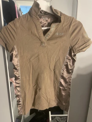 Brun glansig topp från Pikeur - Snygg brun topp från Pikeur med glansiga sidopaneler i satin och korta ärmar. Dekorativa stenar vid knäppningen och loggan på bröstet ger en exklusiv känsla. Hög krage med knapp och figurnära passform. Perfekt för dig som gillar stilrena detaljer.