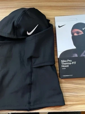 Nike Pro Therma-FIT Hood svart - Nike Pro Therma-FIT Hood i svart, perfekt för fotboll och träning. Tillverkad i syntetmaterial som håller dig varm och torr. Snygg design med vit Nike-logga framtill och tight passform som täcker huvud och hals.