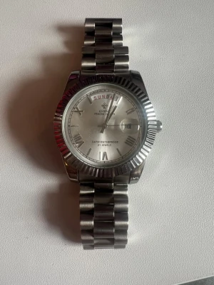 Silverfärgad klocka  - Stilren klocka i silverfärgat metallarmband och rund urtavla. Klockan har romerska siffror, dag- och datumvisning samt räfflad kant runt urtavlan. Perfekt accessoar för en clean look.