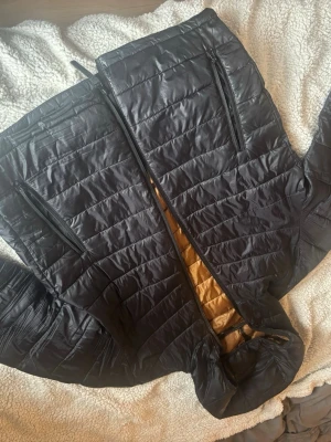 Svart dunväst från Jack & Jones - Snygg svart dunväst från Jack & Jones med guldfärgat foder. Västen har två dragkedjeförsedda fickor framtill och quiltad design. Perfekt för lager på lager och ger en stilren look till din outfit.