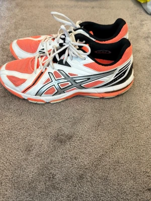 Asics vita och orange sneakers 40 - Snygga sneakers från Asics i vitt och orange med svarta och silvriga detaljer. Skorna har lågt skaft, snörning och meshmaterial för bra andningsförmåga. Perfekta för dig som gillar sportig stil och vill sticka ut med färg.