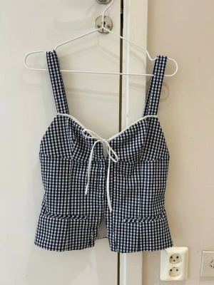  Ny Rutig korsett-topp med snörning - Snygg korsett-topp med breda axelband och vit snörning framtill. Toppens mönster är klassiskt svartvitt rutig och den har en figurnära passform. Perfekt för dig som gillar trendiga och unika plagg med detaljer. Materialet känns som bomull.