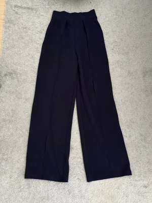 Vida blåa byxor med hög midja - Säljer ett blåa svarta vida byxor med hög midja och snygga pressveck. Byxorna har en stilren look och är tillverkade i ett mjukt, stretchigt material som sitter bekvämt. Perfekta för dig som gillar en avslappnad men trendig stil.