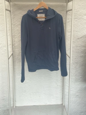 Blå hoodie från Abercrombie & Fitch - Mysig blå hoodie från Abercrombie & Fitch med huva och känguruficka framtill. Liten logga broderad på bröstet och knappar vid halsen. Perfekt för en chill och avslappnad stil. Tillverkad i mjukt bomullsmaterial.