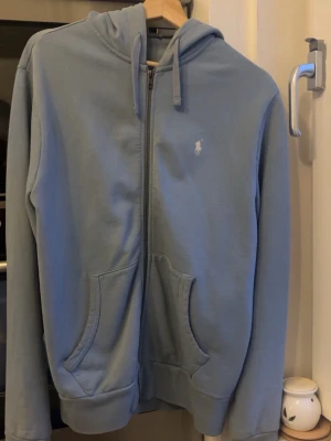 Blå zip-hoodie  Polo Ralph Lauren - Snygg ljusblå hoodie med dragkedja från Polo Ralph Lauren. Klassisk design med huva, två stora fickor fram och logotyp broderad på bröstet. Perfekt för en avslappnad och cool look. Materialet är mjukt och bekvämt.Strl M men passar både S och M. Cond 9/10 köpt från boozt förra året