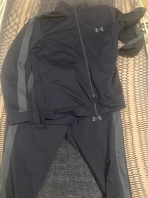 Svart träningsset från Under Armour - Snyggt svart träningsset från Under Armour med gråa ränder längs ärmar och ben. Setet består av en zip-jacka och matchande byxor, båda i mjukt och stretchigt material. Perfekt för gymmet eller sportaktiviteter. Diskret logga på bröstet och höften.