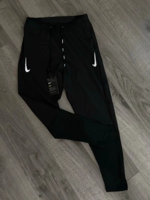 Nike aeroswift S/M - Svarta träningsbyxor från Nike med vita loggor på båda sidor framtill. Byxorna har dragsko i midjan och är tillverkade i ett lätt och snabbtorkande material med Nike Dry-teknologi. Perfekta för träning eller chill. Smal passform och ribbade benslut.