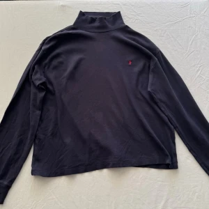 Svart polotröja från Ralph Lauren - Svart långärmad polotröja från Ralph Lauren med liten röd logga broderad på bröstet. Tröjan har en hög krage och är tillverkad i mjukt material som känns skönt mot huden. Perfekt för lager-på-lager och stilrena outfits.