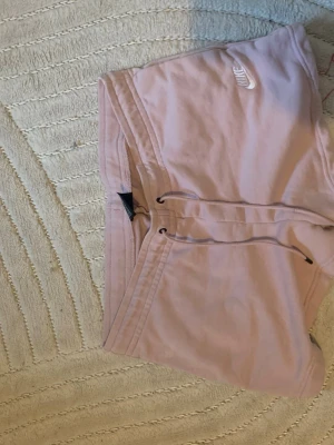 Ljusrosa shorts från Nike med snörning - Säljer ett par ljusrosa shorts från Nike med vit logga på ena benet. Shortsen har snörning i midjan och är tillverkade i mjuk bomull, perfekta för chill dagar eller träning. Klassisk passform och enkel design.