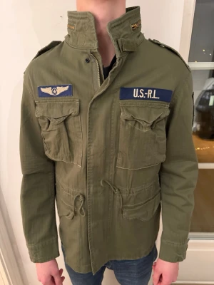 Grön militärinspirerad jacka U.S.RL - Snygg militärgrön jacka från U.S.RL med broderade patchar och detaljer. Jackan har fyra stora fickor framtill, snörning i midjan och dragkedja. Axelpartierna har dekorativa märken och jackan är tillverkad i bomull. Perfekt för en cool och avslappnad stil.