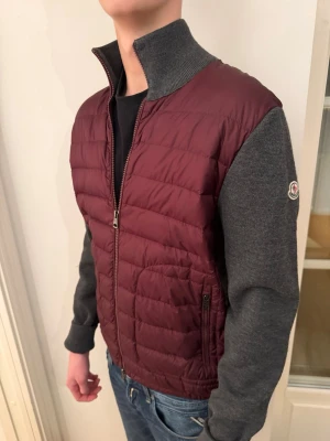 Moncler vinröd och grå pufferjacka - Snygg pufferjacka från Moncler med vinröd, quiltad front och grå stickade ärmar och rygg. Jackan har hög krage, dragkedja framtill och Moncler-logga på ärmen. Perfekt för dig som vill ha en modern och sportig look.