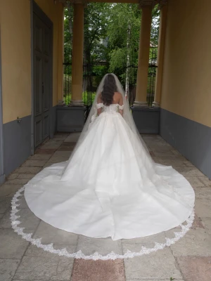 Vit lång brudklänning med spetsdetaljer - Elegant vit brudklänning med offshoulder och vacker spets upptill. Klänningen har en vid kjol med långt släp och fina spetsapplikationer längs kanten. Materialet är troligtvis en blandning av satin och spets, vilket ger en lyxig känsla. Perfekt för dig som vill ha en klassisk och romantisk look.