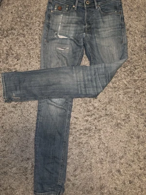 Blå slitna slim jeans - Säljer ett par blå slim jeans med slitningar och distressed detaljer på framsidan. Jeansen har klassisk femficksdesign, normalhög midja och är tillverkade i denim. Perfekta för en avslappnad och trendig look, Jeansen har tre defekter en lite större vid högra framfickan men kan sys ihop och två mindre en brevid gylfen och en vid nedre benet, 