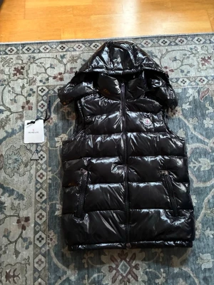 Svart Moncler väst - Snygg svart dunväst från Moncler med glansig finish och avtagbar huva. Västen har två dragkedjeförsedda fickor fram och klassisk Moncler-logga på bröstet. Perfekt för lager på lager och streetstyle. Materialet är dunfyllning och yttertyget är blankt. Västen är i fint skick men används inte längre, den har även avtagbar luva
