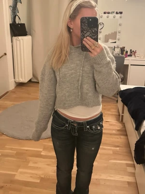 Grå croppad stickad tröja - Säljer en grå croppad stickad tröja med långa ärmar och ribbade muddar. Tröjan har en avslappnad passform och är perfekt för lager-på-lager. Mjuk och mysig känsla, passar dig som gillar trendiga och bekväma plagg. Har även en luva.